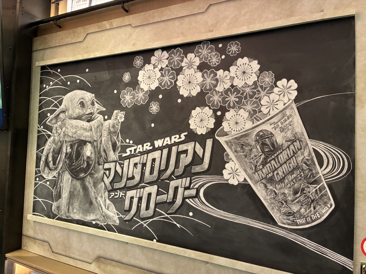 黒板にチョークで描かれたグローグーのイラストと映画タイトル。紙のカップから花びらが溢れ出している