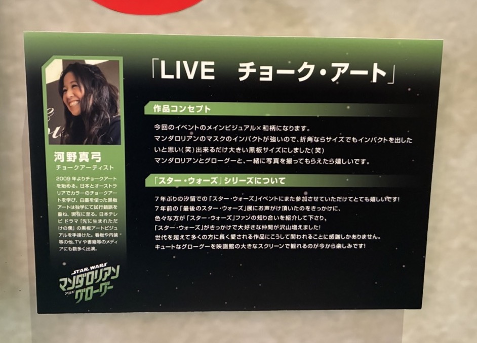 チョークアートの作者、河野真弓さんの紹介パネル。以下コンセプト「今回のイベントのメインビジュアル✕和柄になります。マンダロリアンのマスクのインパクトが強いので、折角ならサイズでもインパクトを出したいと思い（笑）出来るだけ大きい黒板サイズにしました（笑）マンダロリアンとグローグーと、一緒に写真を撮ってもらえたら嬉しいです。」以下コメント「7年ぶりの汐留での「スター・ウォーズ」イベントにまた参加させていただけてとても嬉しいです！7年前の「最後のスター・ウォーズ」展にお声がけ頂いたのをきっかけに、色々な方が「スター・ウォーズ」ファンの知り合いを紹介して下さり、「スター・ウォーズ」がきっかけで大好きな仲間が沢山増えました！世代を超えて多くの方に長く愛される作品にこうして関われることに感謝しかありません。キュートなグローグーを映画館の大きなスクリーンで観れるのが今から楽しみです！」以下プロフィール「2009年よりチョークアートを始める。日本とオーストラリアでカラーのチョークアートを学び、白墨を使った黒板アートは独学にて試行錯誤を重ね、現在に至る。日本テレビドラマ「先に生まれただけの僕」の黒板アートビジュアルを手掛けた。看板や内装・等の他、TVや書籍等のメディアにも数多く出演。」