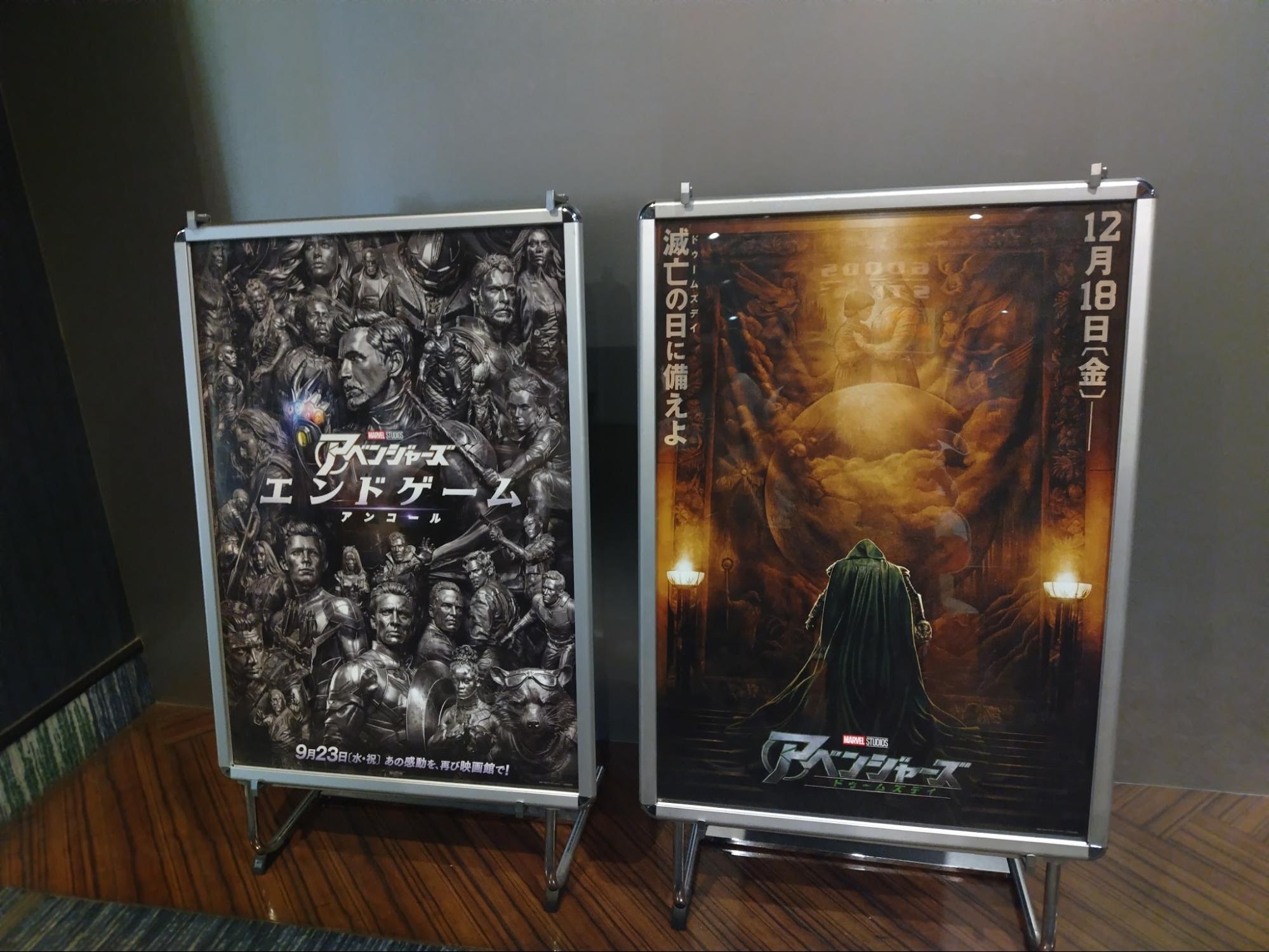 映画『アベンジャーズ／エンドゲーム：アンコール』のポスターと、映画『アベンジャーズ／ドゥームズデイ』のキービジュアルが並んで展示されている。『ドゥームズデイ』のキービジュアルには、緑のフードを被った人物の後ろ姿が描かれ、12月18日（金）公開と書かれている